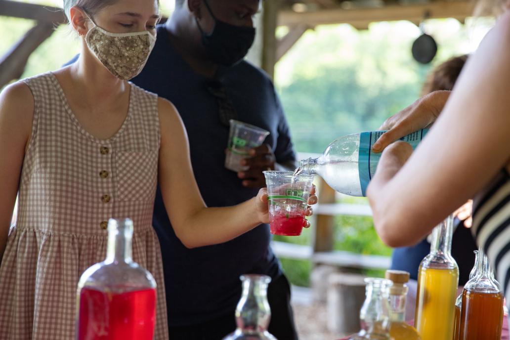 A participant hold out a cup with red syrup and ice, seltzer is poured over top.