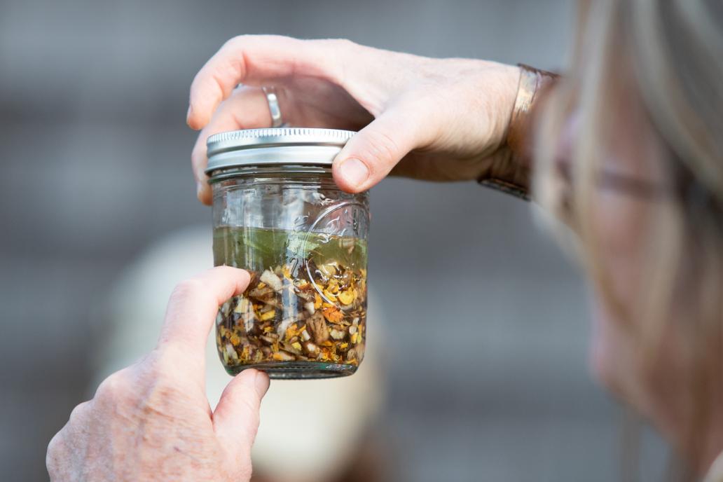 A person holds up a lidded glass jar, full of herbal ingredients and steeped in a clear liquid.