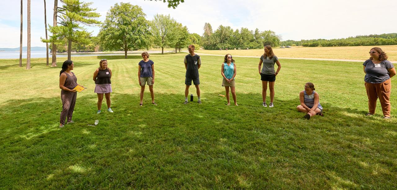 A semicircle of eight educators stand in a shaded grassy field in summertime, with Lake Champlain and the Adirondack Mountains in the distance.