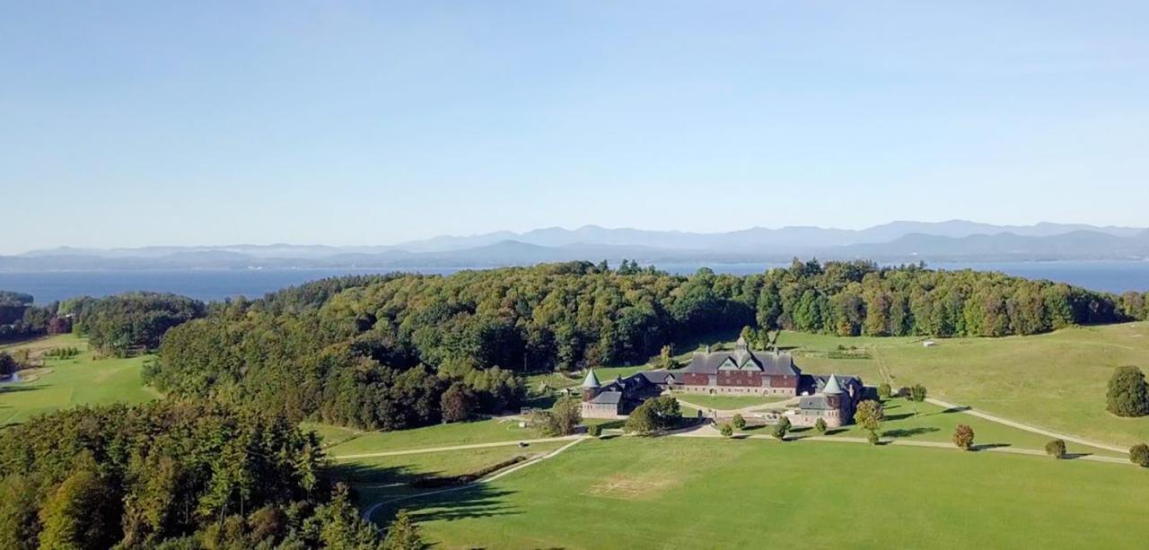 An aerial view of a sweeping Vermont landscape. A large barn stands at center surrounded by rolling green pastures and tree-covered hills. Lake Champlain and the Adirondack mountains stand in the distance.