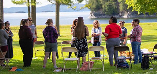 Two dozen educators stand in a circle on a grassy lawn engaged in a team building activity. Lake and mountains are seen in distance.