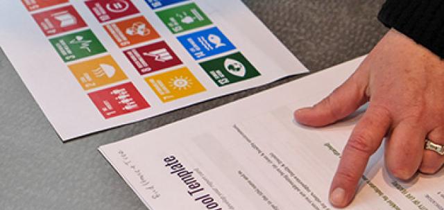 Overhead view of a table. An educator points on a worksheet with colorful Sustainable Development Goals printed on paper.