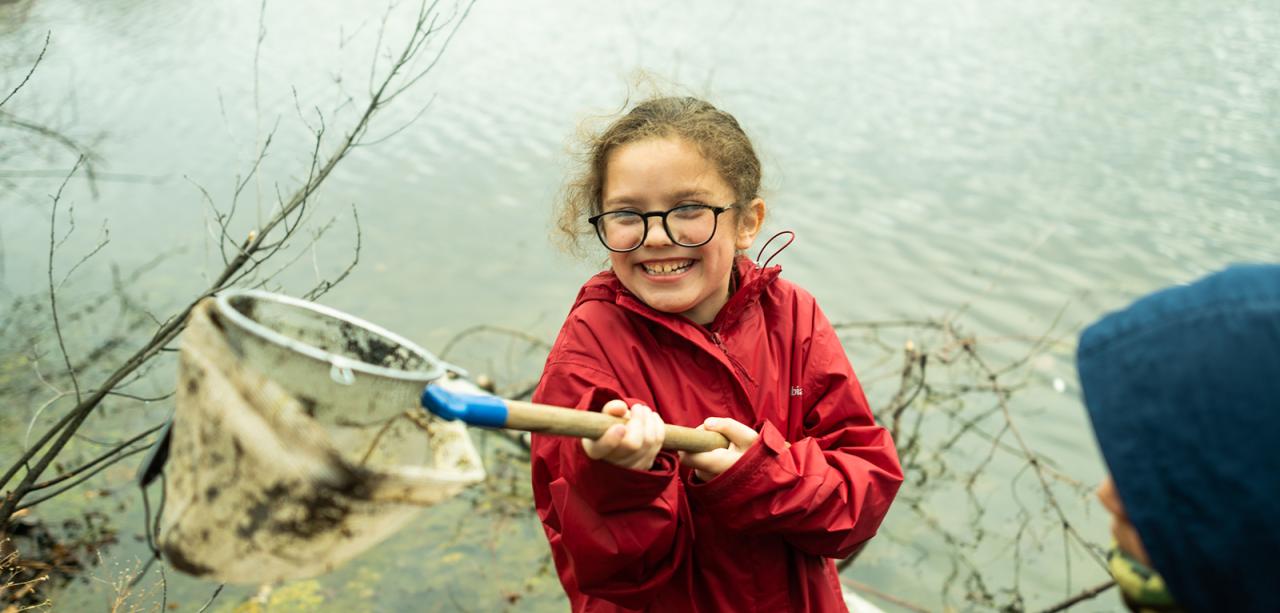 An elementary aged student holds a net and smiles in front of a pond in springtime. The net is full of leaf debris.
