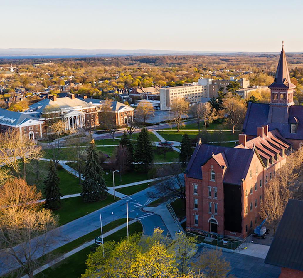 Aerial view of the University of Vermont campus in springtime. Large towering buildings stand in foreground with grassy school green in middleground. Rolling hills stand in background.